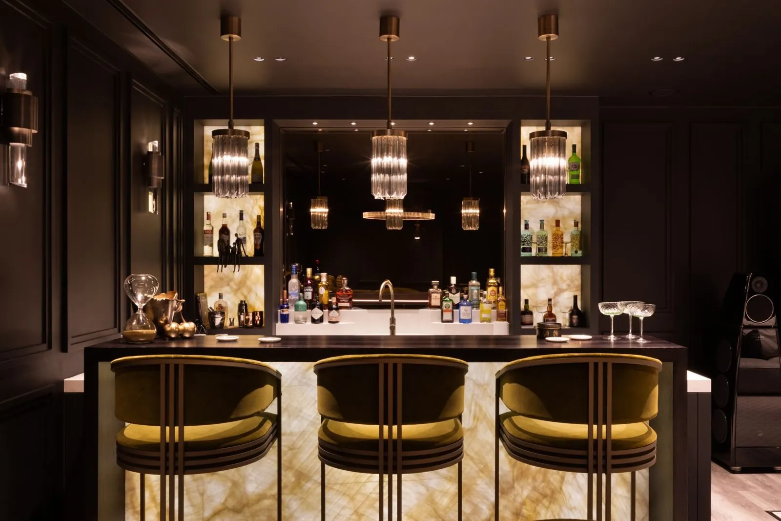 A private bar with a backlit onyx counter, brass-framed stools and crystal pendants